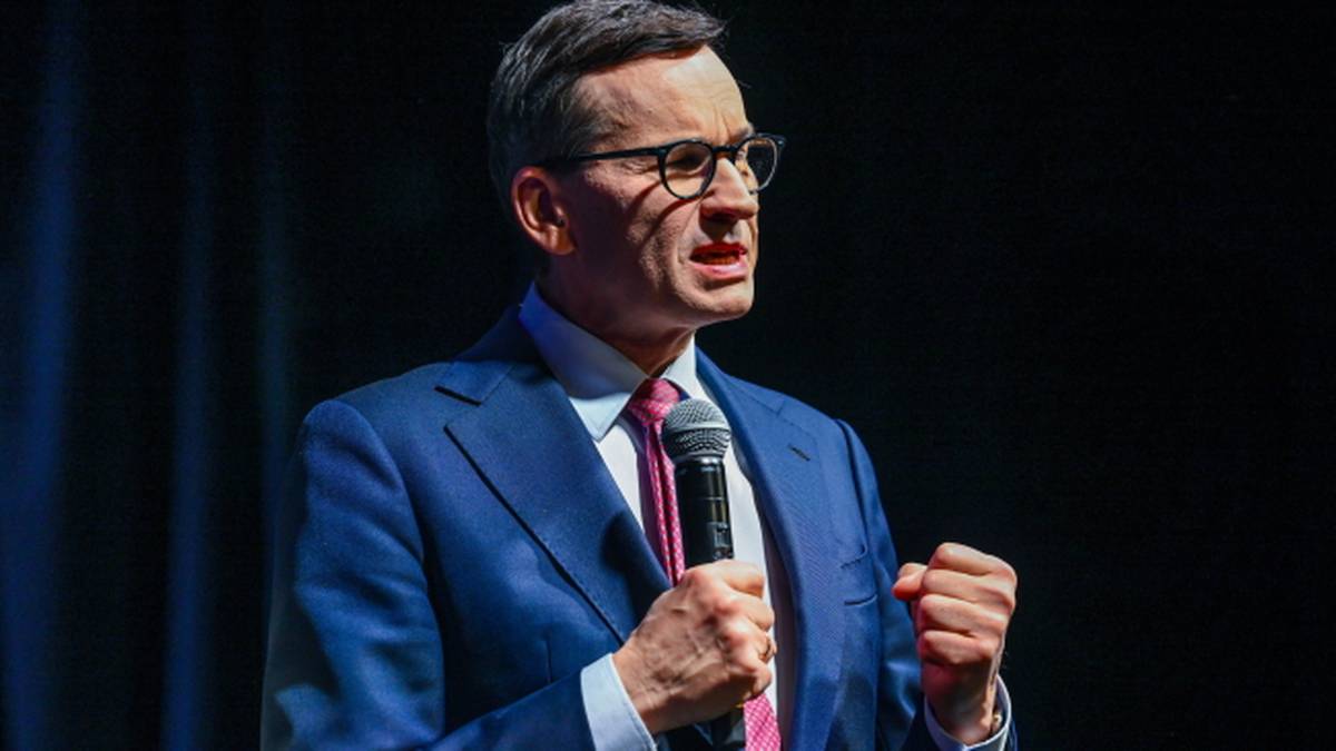 Mężczyzna w niebieskim garniturze i okularach przemawia do mikrofonu.