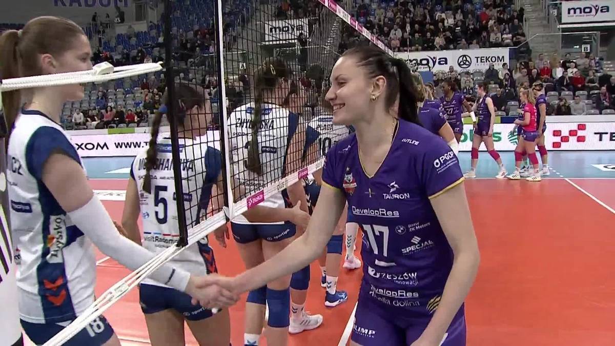 Dwie siatkarki ściskają sobie dłonie przy siatce, obie w strojach sportowych.