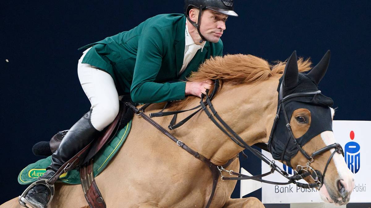 Jeździec w zielonej marynarce i białych bryczesach dosiada jasnobrązowego konia, pokonując przeszkodę. Obydwaj skupieni są na zadaniu.