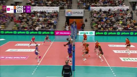 Mecz siatk&oacute;wki między drużynami Developres Rzesz&oacute;w a UNI Opole. Zawodniczka Developres Rzesz&oacute;w skacze do ataku nad siatką, podczas gdy zawodniczki UNI Opole pr&oacute;bują ją zablokować. Na tablicy wynik&oacute;w widnieje wynik 19:21 dla Developres Rzesz&oacute;w.