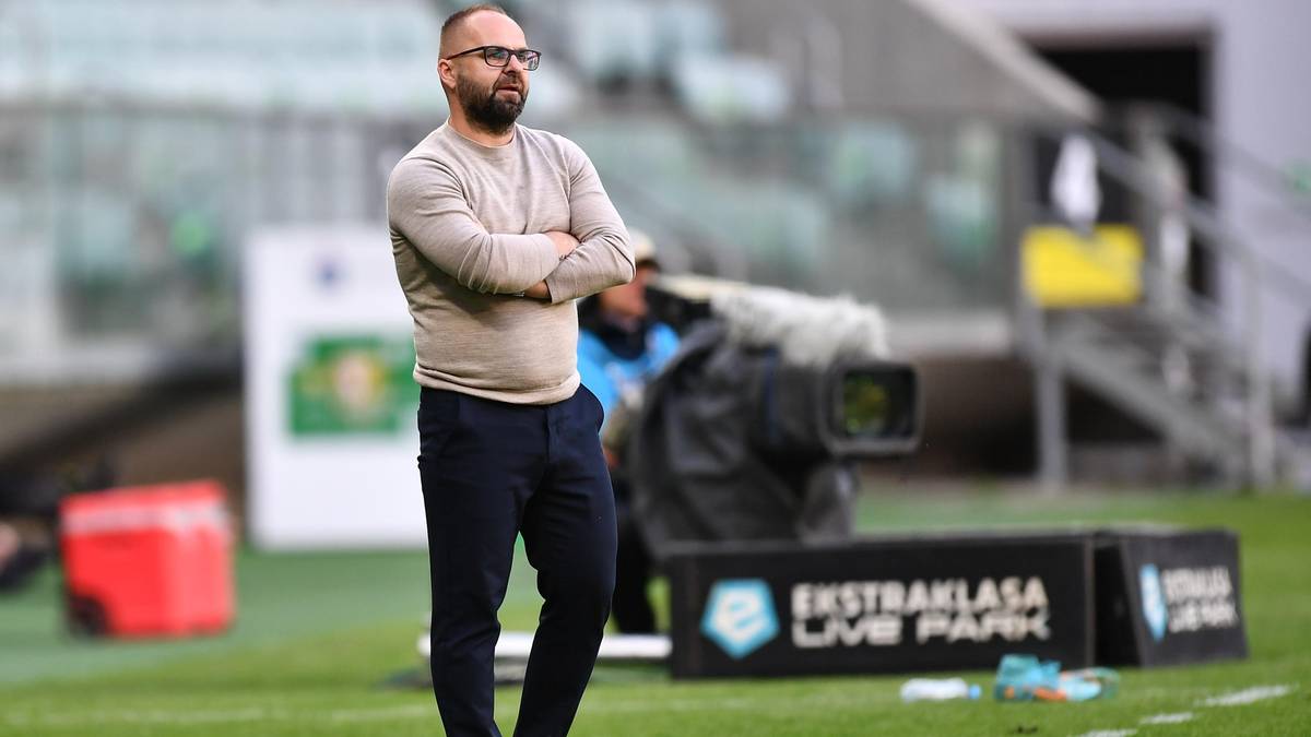 Trener piłkarski w okularach z założonymi rękami na piersi stoi na boisku.