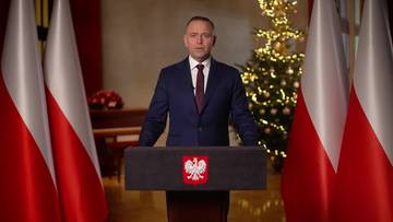 Prezydent Karol Nawrocki stoi za m&oacute;wnicą z godłem Polski, w tle trzy flagi Polski i choinka.