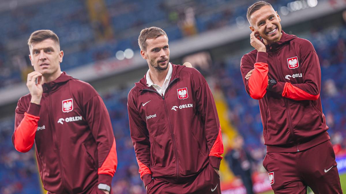 Trzech piłkarzy reprezentacji Polski w bordowych dresach z orłem na piersi, stojący na murawie stadionu.