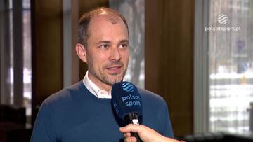 Mężczyzna w niebieskim swetrze z białym kołnierzykiem, trzymający mikrofon z logo Polsat Sport.