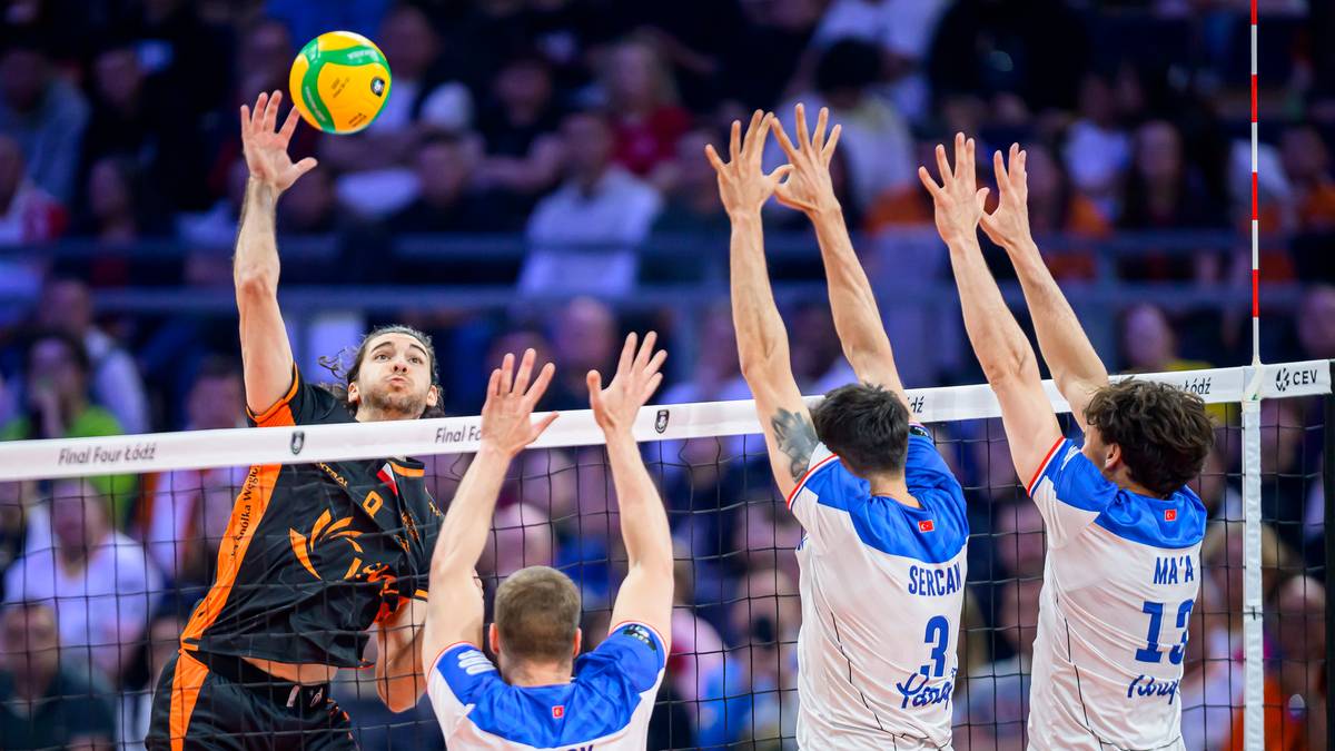 Siatkarz w czarno-pomarańczowej koszulce odbija piłkę nad siatką, a czterech przeciwników w niebiesko-białych strojach próbuje ją zablokować.