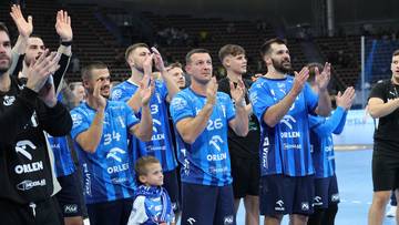 Grupa mężczyzn w niebieskich strojach sportowych, niektórzy z numerami na koszulkach, klaszcze i patrzy w górę. W tle widać trybuny z widzami. Na pierwszym planie stoi chłopiec w niebieskim stroju.