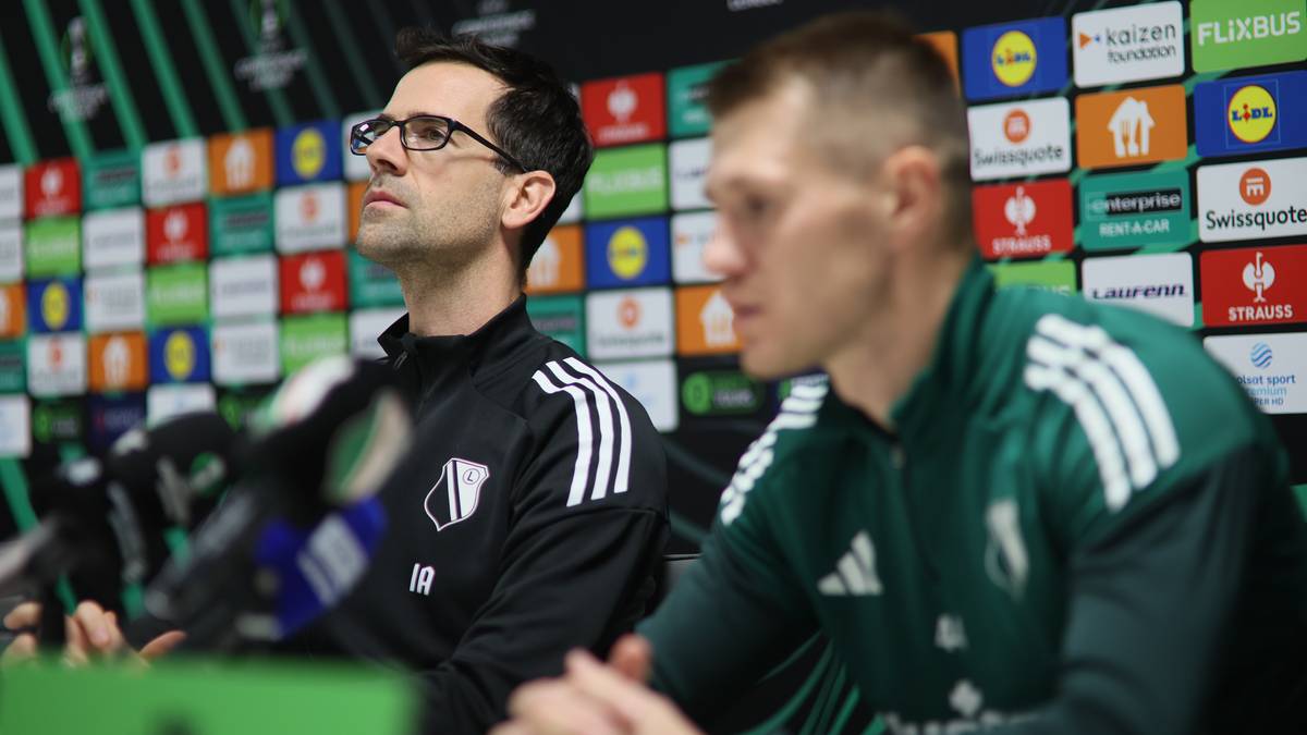 Dwóch mężczyzn, prawdopodobnie trenerzy lub przedstawiciele klubu, siedzi za stołem podczas konferencji prasowej. Jeden z nich ma okulary, drugi patrzy w bok. W tle widoczna jest ściana z kolorowymi logotypami sponsorów.