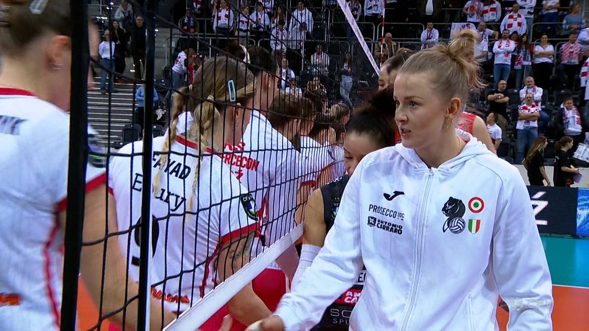Siatkarka w białej bluzie z emblematami wychodzi zza siatki podczas meczu.