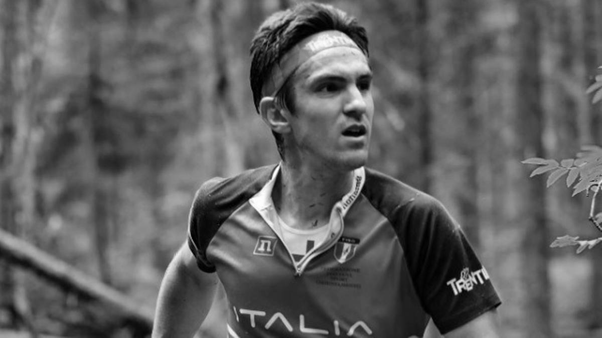 Czarno-białe zdjęcie młodych mężczyzny w stroju sportowym, biegnącego w lesie. Ma na głowie opaskę. Widoczna jest na jego twarzy determinacja.