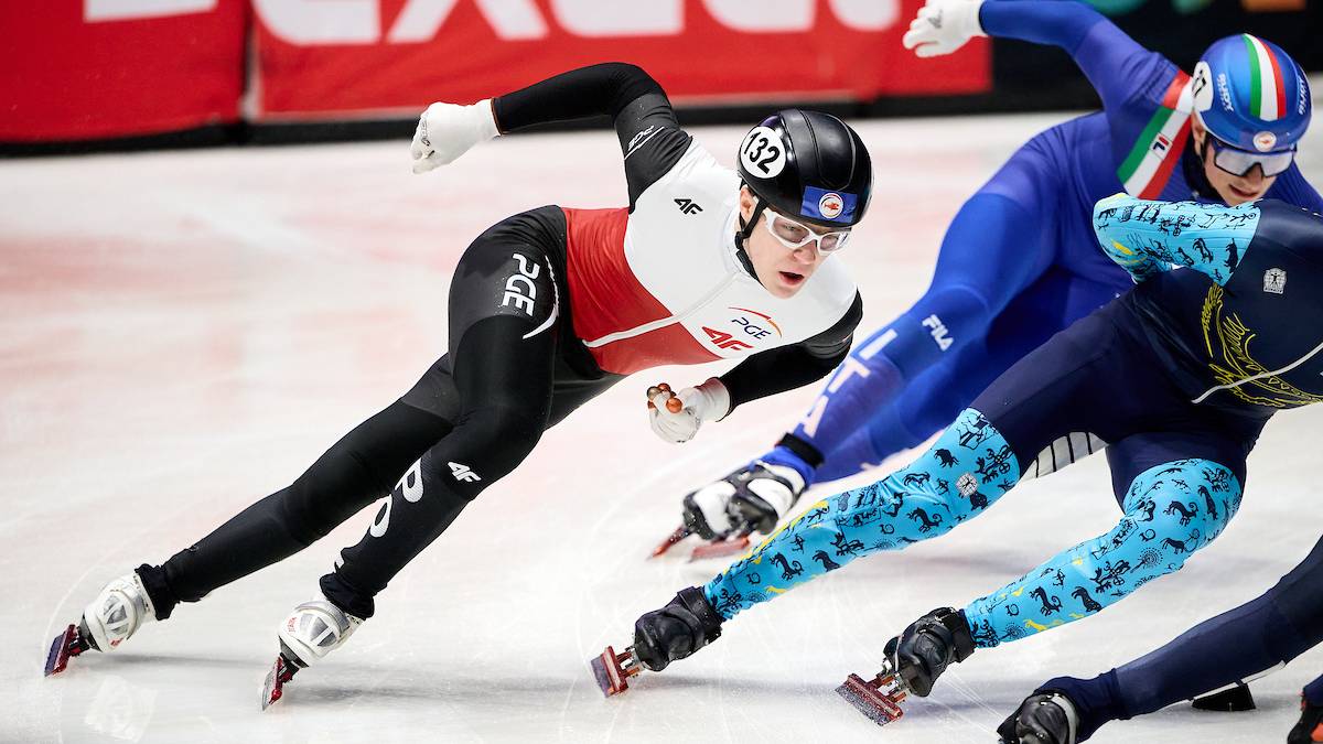 Trzy zawodniczki w strojach sportowych jadą na łyżwach po torze lodowym podczas zawodów short tracku. Jedna z nich, ubrana w biało-czerwony strój z numerem "32" na kasku, jest na pierwszym planie, w pozycji pochylonej i dynamicznej. Po jej prawej stronie jadą dwie inne zawodniczki, jedna w niebieskim stroju, druga w ciemnym stroju z kolorowymi wzorami na nogawkach.