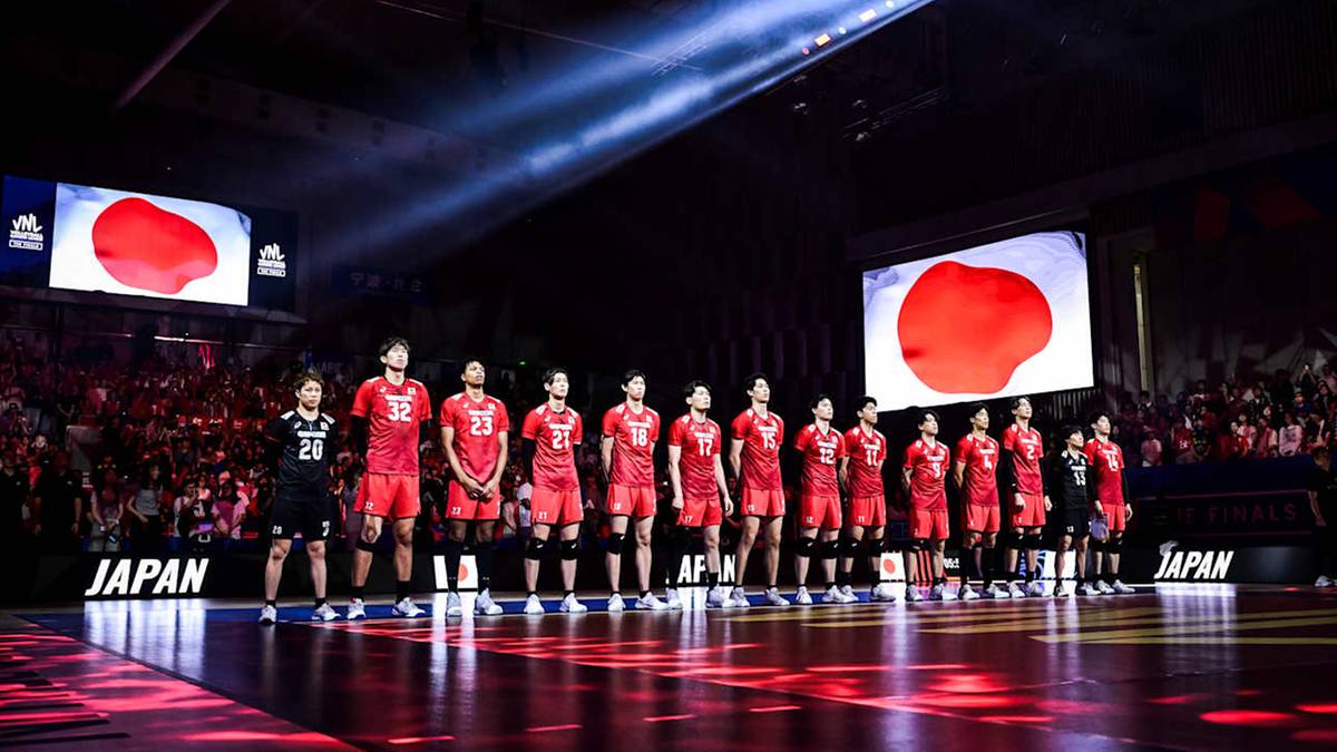 Drużyna siatkarzy Japonii pozuje do zdjęcia w strojach sportowych z czerwonymi elementami, stojąc przed dużym ekranem wyświetlającym logo w kształcie czerwonej kuli.