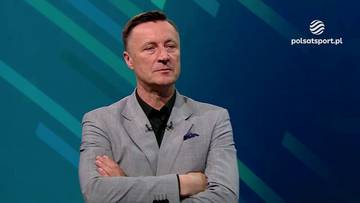 Tomasz Hajto w jasnej marynarce i czarnej koszuli, z rękami skrzyżowanymi na piersi, patrzy w bok. W tle znajduje się niebiesko-zielone tło z logo Polsat Sport.
