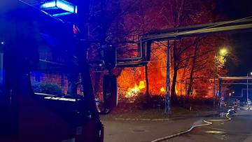 Nocny pożar na terenie zakładu przemysłowego, widoczne płomienie i wóz strażacki z wężem gaśniczym.