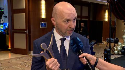 Dariusz Krysiak w garniturze trzyma statuetkę i udziela wywiadu przed mikrofonem.