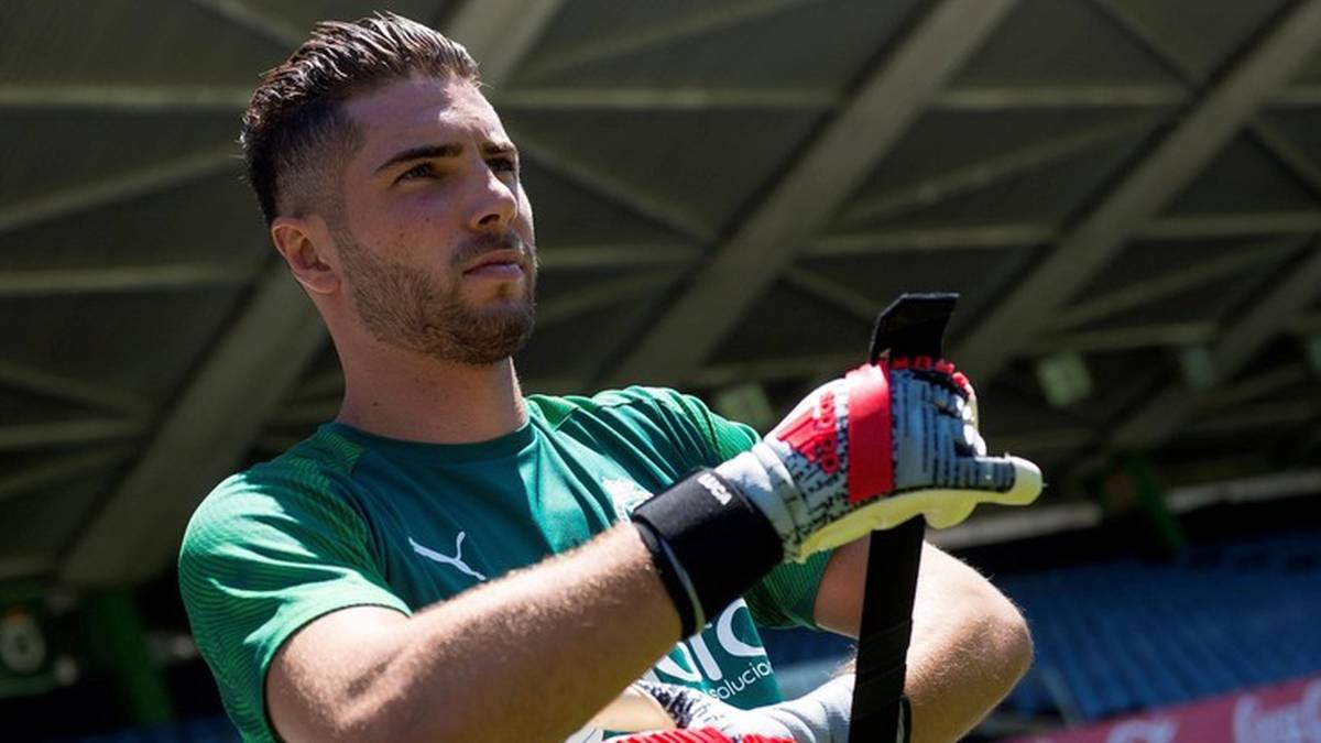 Młody mężczyzna w zielonej koszulce bramkarskiej i rękawicach, trzymający kij hokejowy.