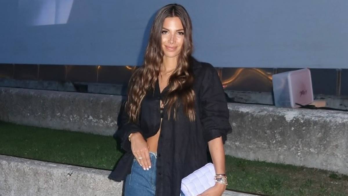 Młoda kobieta w czarnej, rozpiętej koszuli i dżinsach stoi z dokumentami w ręku. Uśmiecha się lekko do aparatu.