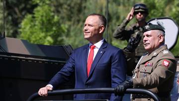 Mężczyzna w garniturze z czerwoną krawatem stoi obok żołnierza w mundurze na tle zieleni.