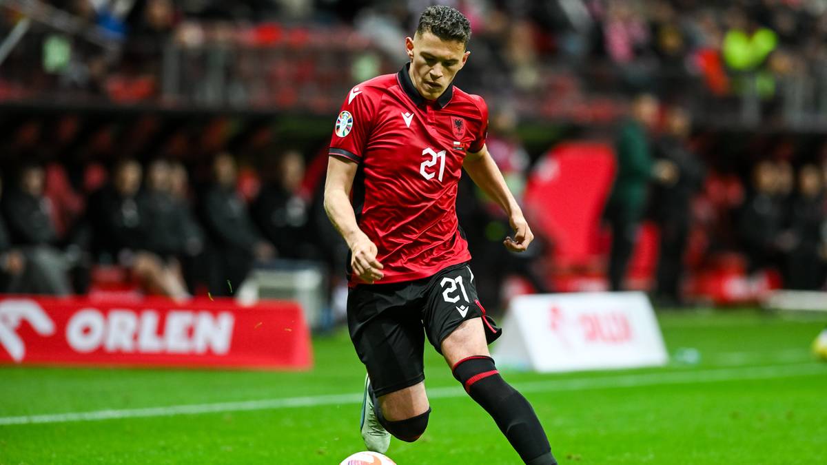 Piłkarz reprezentacji Albanii w czerwonej koszulce z numerem 21 prowadzi piłkę na boisku stadionu.