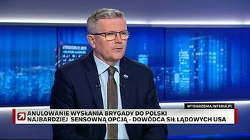 Mężczyzna w średnim wieku, ubrany w garnitur i krawat, stoi przed ekranem z niebieskim tłem przedstawiającym nocną panoramę miasta. Na ekranie widoczny jest r&oacute;wnież napis "WYDARZENIA.INTERIA.PL" oraz "ANULOWANIE WYSŁANIA BRYGADY DO POLSKI NAJBARDZIEJ SENSOWNĄ OPCJĄ - DOW&Oacute;DZTWA LĄDOWYCH USA".