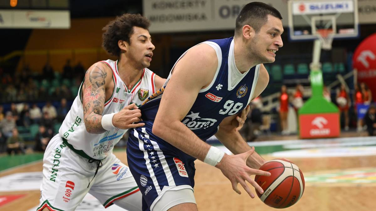 Dwóch koszykarzy na boisku. Zawodnik w białej koszulce broni zawodnika w granatowej koszulce, który prowadzi piłkę.