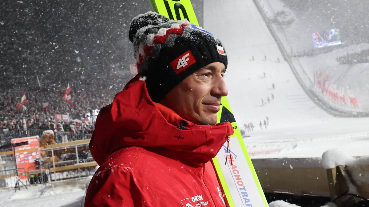 Skoczek narciarski w czerwonej kurtce trzyma narty przed skocznią.