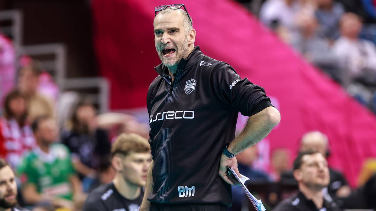 Trener siatkówki mężczyzna w czarnej bluzie z krótkim rękawem, z mikrofonem w dłoni, krzyczy do swoich zawodników na boisku.