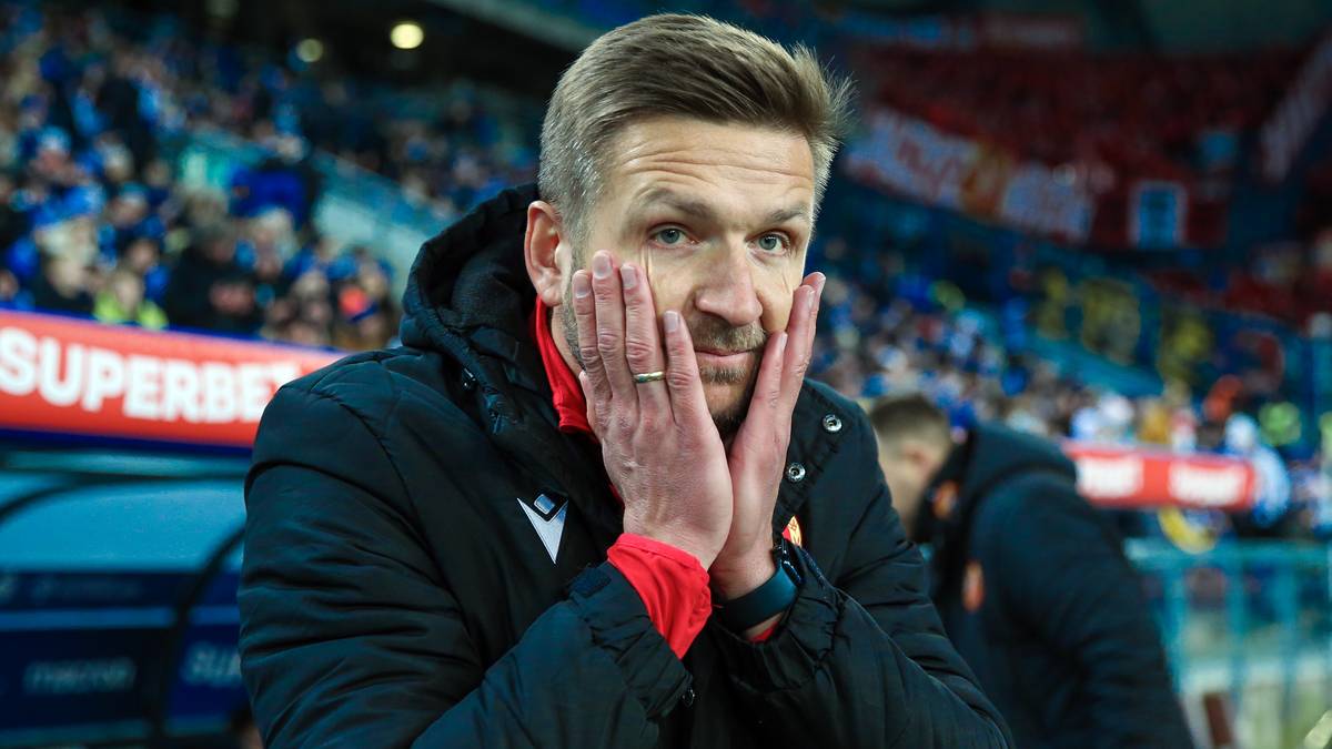 Trener piłkarski w czarnej kurtce, z rękami przy twarzy, wyrażający zmartwienie lub frustrację. W tle trybuny stadionu.