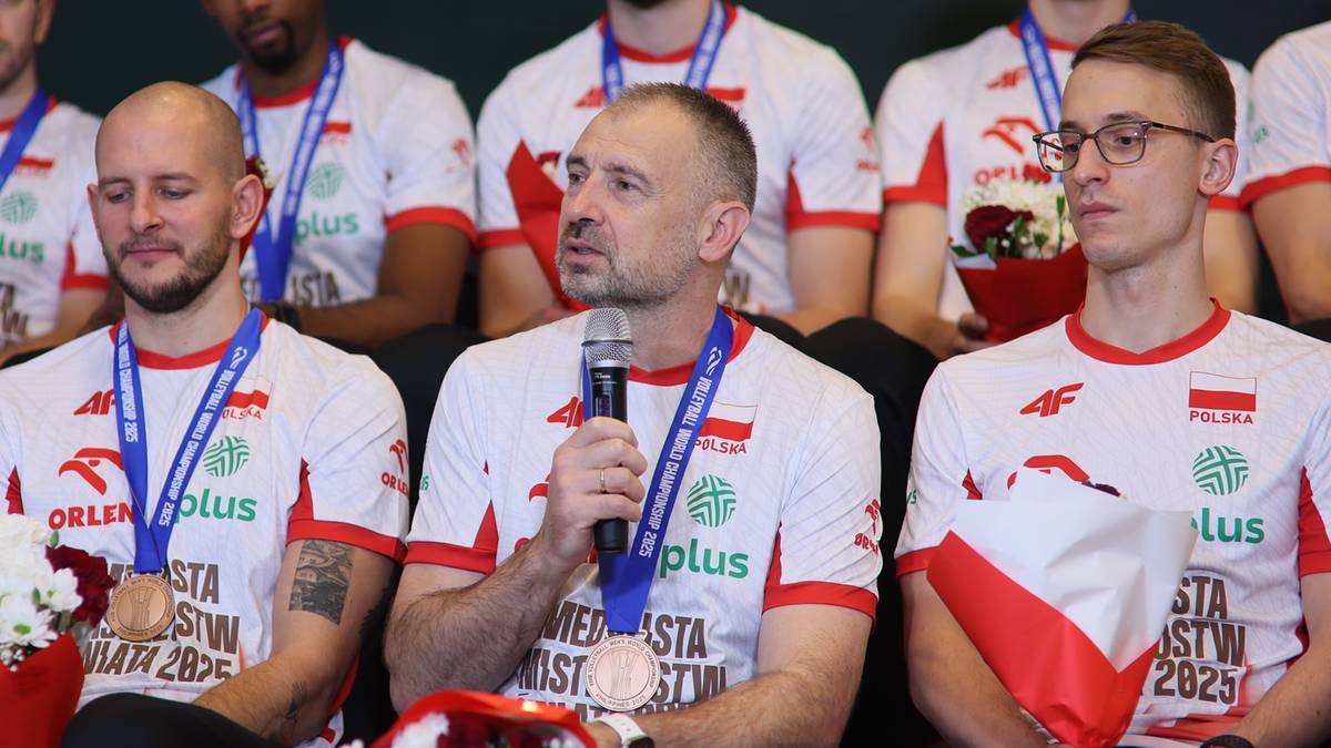 Trzech mężczyzn w strojach sportowych siedzi w rzędzie. Środkowy mężczyzna przemawia do mikrofonu, a pozostali patrzą przed siebie. Dwóch z nich ma na szyi niebieskie szarfy, a jeden z nich trzyma medal.