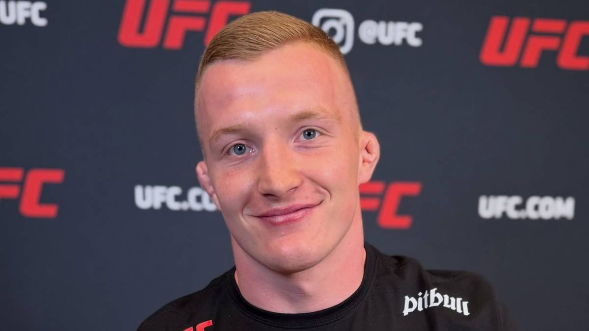 Młody mężczyzna o jasnych włosach i niebieskich oczach, uśmiecha się. Za nim widnieje ciemne tło z logo UFC.
