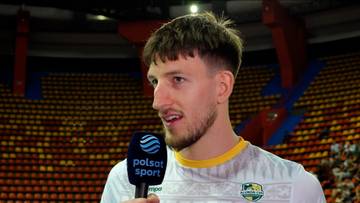 Mężczyzna w koszulce sportowej stoi przed widownią na stadionie i udziela wywiadu do mikrofonu Polsat Sport.