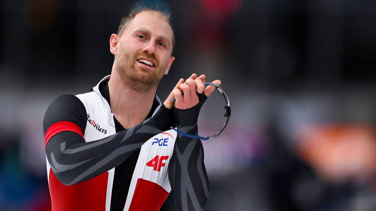 Łyżwiarz szybki w czerwono-czarnym stroju trzyma okulary w dłoni, patrząc w stronę kamery z lekkim uśmiechem.