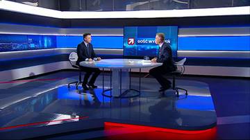 Dw&oacute;ch mężczyzn w garniturach rozmawia w studiu telewizyjnym, siedząc naprzeciwko siebie przy okrągłym stole. W tle widoczny jest ekran z logo stacji.