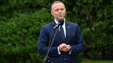 Mężczyzna w garniturze i krawacie przemawia do mikrofonu na tle zielonych krzew&oacute;w.