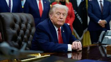 Prezydent USA Donald Trump siedzi przy biurku w Gabinecie Owalnym, otoczony doradcami.