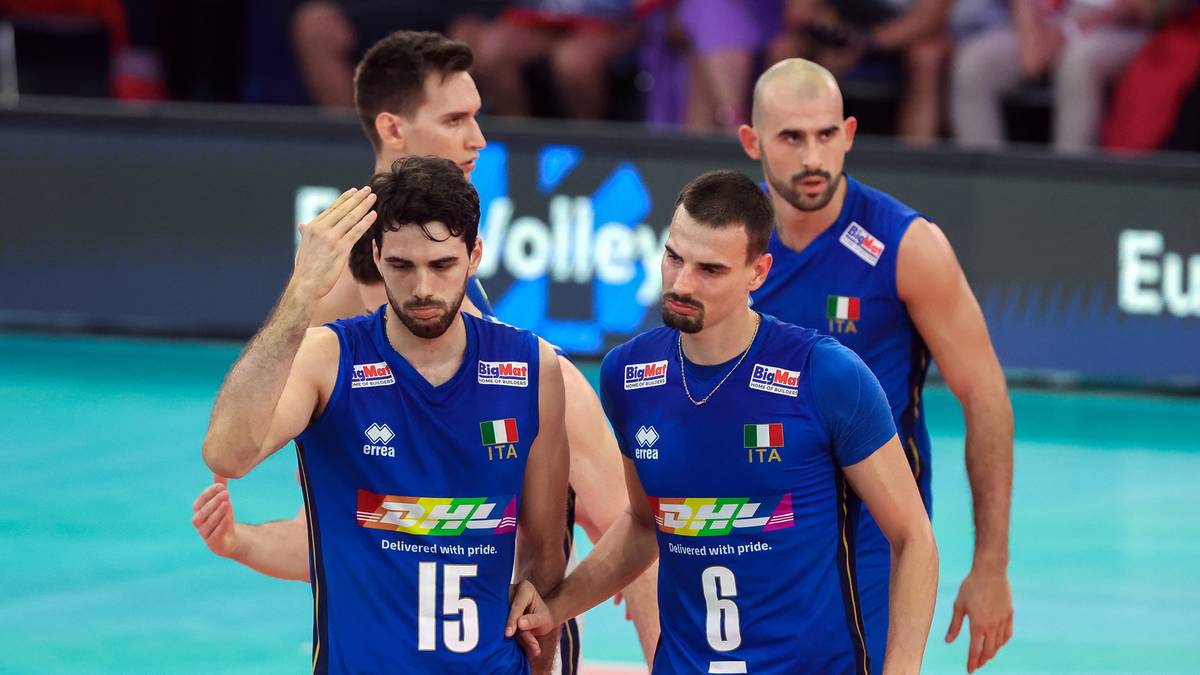 Siatkarz w niebieskiej koszulce z numerem 15, z opaską na ramieniu, zasłania czoło. Obok stoi inny zawodnik z numerem 6. W tle widoczni są inni gracze w podobnych strojach.