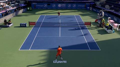 Kort tenisowy podczas meczu z dwoma zawodnikami. Jeden z nich jest bliżej kamery, drugi na przeciwległej stronie. Widoczni są widzowie na trybunach i reklamy.