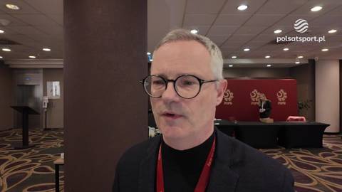 Mężczyzna w okularach i czarnym swetrze z golfem, m&oacute;wiący do kamery. Tło to sala konferencyjna z czerwonymi elementami dekoracyjnymi.