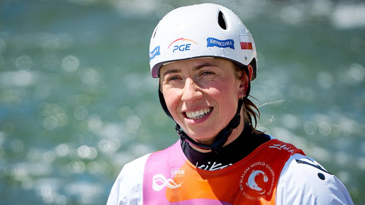 Kobieta w białym kasku narciarskim z logo PGE i niebieskimi napisami, uśmiechnięta, w stroju sportowym do kajakarstwa, na tle rozmytej wody.