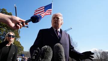 Donald Trump przemawia do mikrofon&oacute;w pod amerykańską flagą.