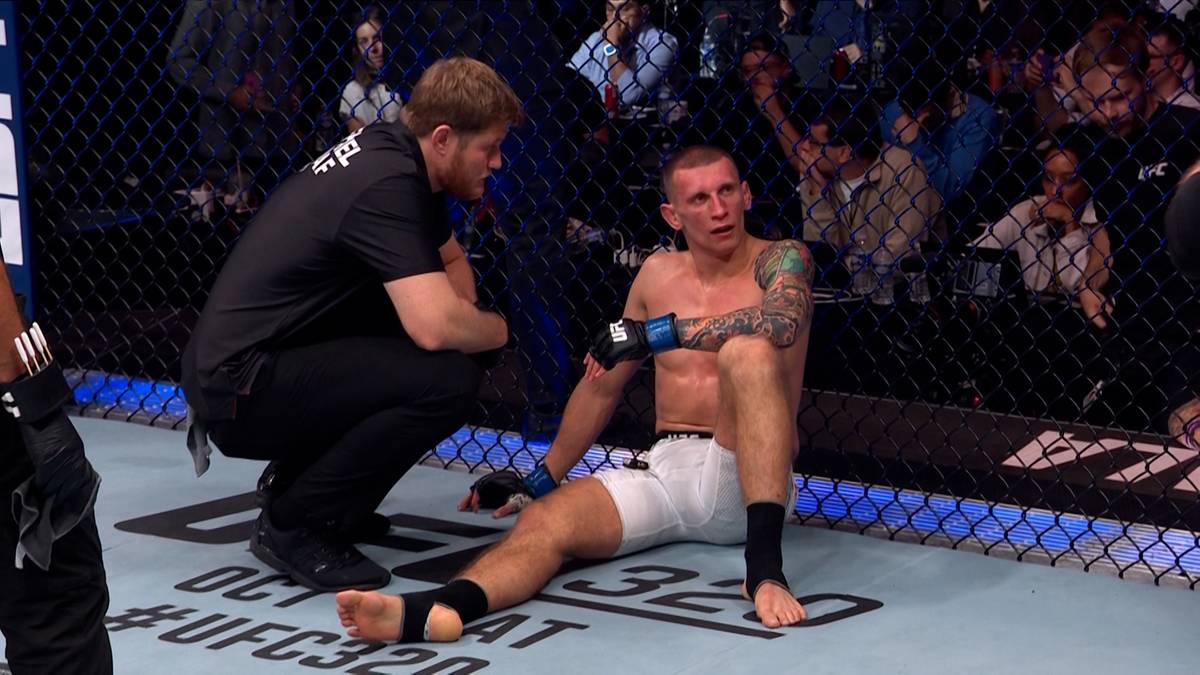 Zawodniczka siedzi na macie oktagonu, pochylony nad nim trener podaje mu ręcznik i mówi coś do niego.