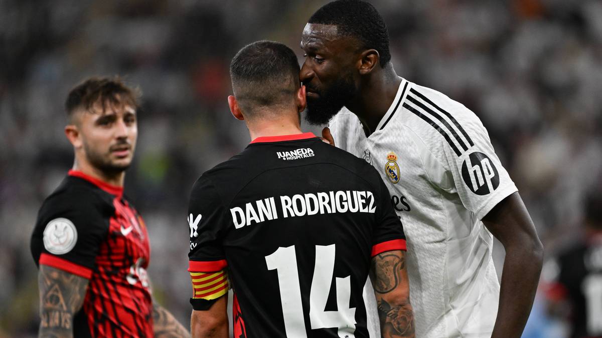 Piłkarze w akcji na boisku podczas meczu piłki nożnej. Jeden z nich, ubrany w białą koszulkę Realu Madryt, pochyla się i rozmawia z innym zawodnikiem w czarnej koszulce.