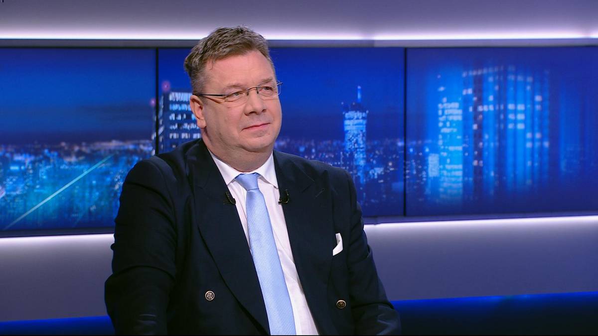 Mężczyzna w okularach, marynarce i krawacie siedzi przed ekranem z widokiem na nocne miasto.