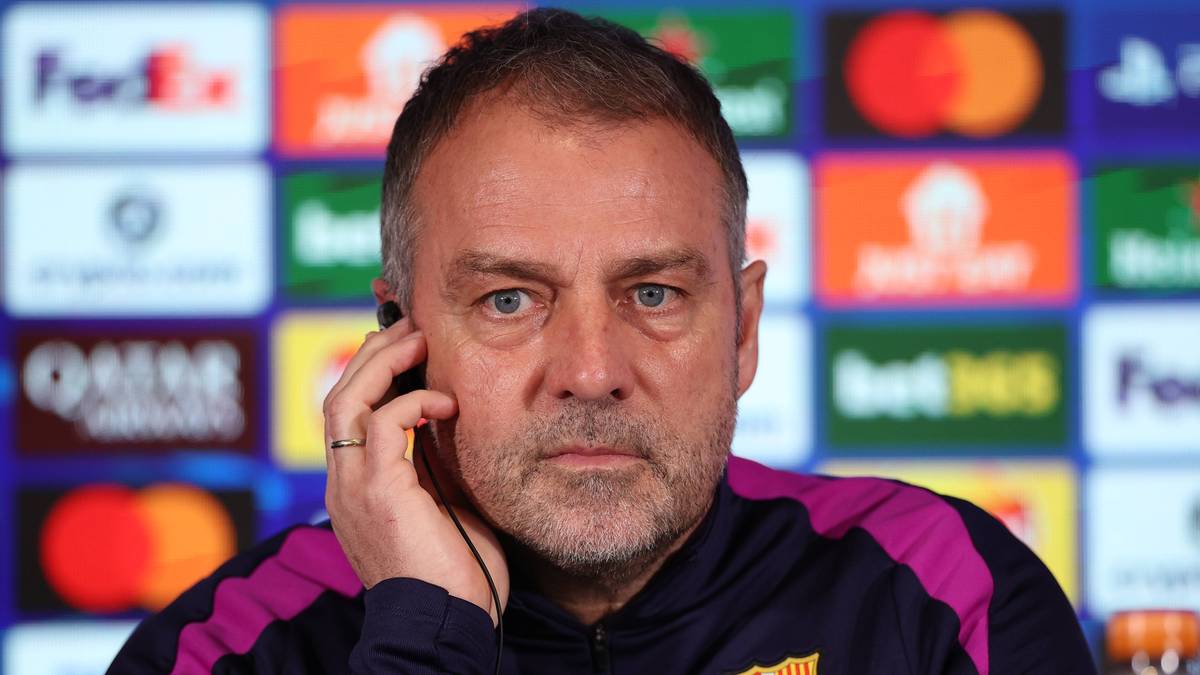 Portret mężczyzny w średnim wieku z kr&oacute;tkimi, siwiejącymi włosami i kilkudniowym zarostem, patrzącego prosto przed siebie z poważnym wyrazem twarzy. Ma na sobie granatową bluzę sportową z fioletowymi i r&oacute;żowymi pasami na ramionach. W uchu ma słuchawkę, a prawą ręką dotyka swojego policzka. W tle widoczny jest panel z logo Ligii Mistrz&oacute;w UEFA oraz nazwami sponsor&oacute;w.