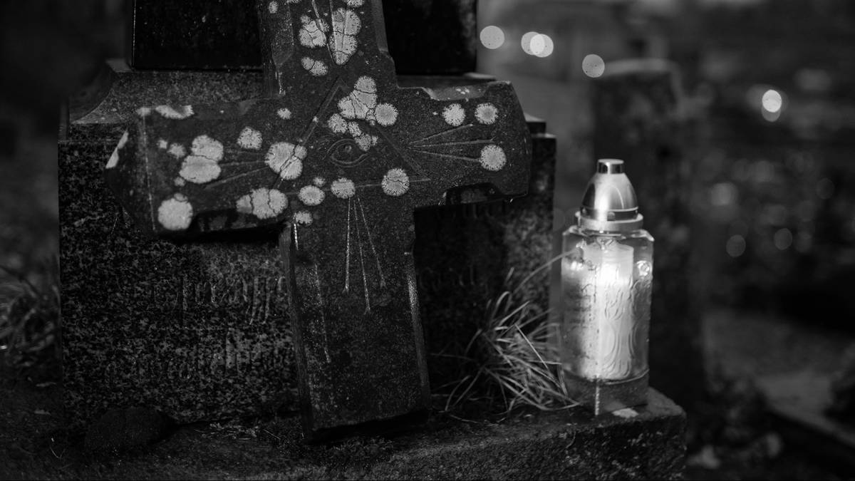 Kamienny krzyż nagrobny z plamami, obok którego płonie znicz. Tło jest ciemne z rozmazanymi światłami.