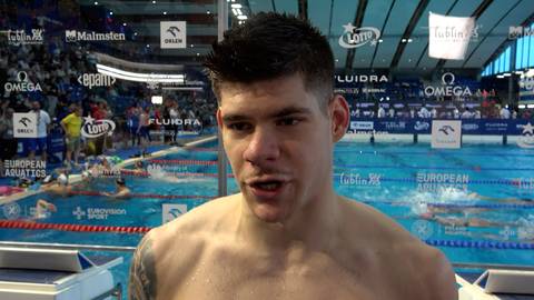 Młody mężczyzna, pływak, w zbliżeniu na tle basenu pływackiego z torami i trybunami.