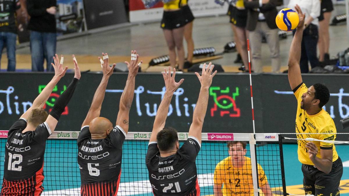 Mecz siatk&oacute;wki, gracz w ż&oacute;łtym stroju atakuje, czterech graczy w czarnych strojach pr&oacute;buje zablokować.