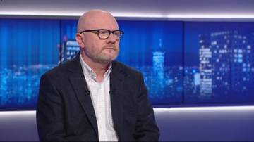 Mężczyzna z łysą głową i brodą w okularach, ubrany w marynarkę i białą koszulę, siedzi przed ekranami z nocnym widokiem na miasto.