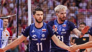 Czterech siatkarzy w granatowych strojach świętuje zwycięstwo, podając sobie ręce, z siatką i kibicami w tle.