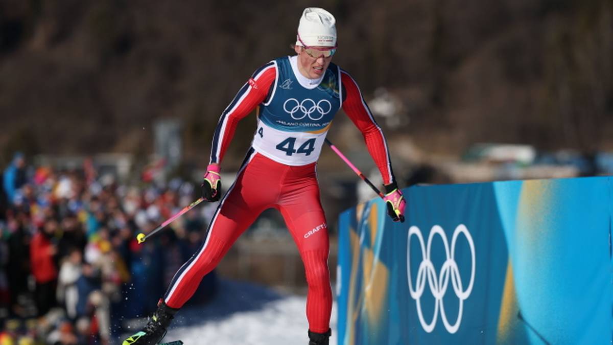 Narciarz biegowy w czerwono-białym stroju z numerem 44 na piersi, biegnie na nartach po śniegu, obok niebieska bandyta z logo igrzysk olimpijskich.