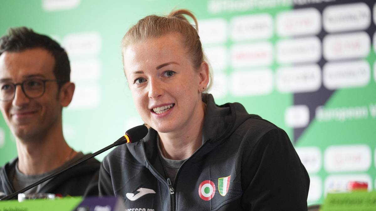 Zbliżenie na uśmiechniętą kobietę z blond włosami spiętymi w kucyk, siedzącą przy mikrofonie. Na jej ramieniu widoczne logo drużyny z flagą Węgier. W tle widać mężczyznę.